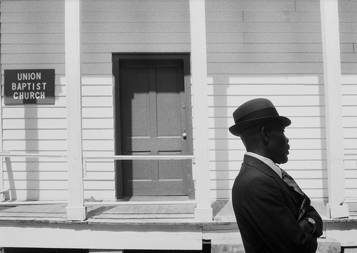 photograph of man in suit and hat walking past front of white church